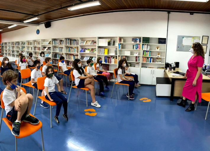 Foto da jornalista Bruna durante conversa com os estudantes do Colégio Ofélia Fonseca. Bruna está na frente das crianças, que estão sentadas em cadeiras, na biblioteca.